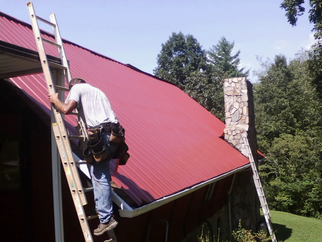 Roofer on a ladder beside a red metal roof and stone chimney — Soffit & Fascia Repair by Roofing Brothers in South Easton